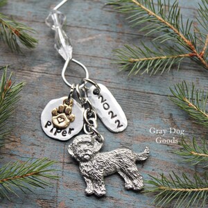 May include: A silver dog charm keychain with a small charm that says "Piper" and a paw print. The keychain also has a charm that says "2022". The background includes pine needles and the text "Gray Dog Goods".