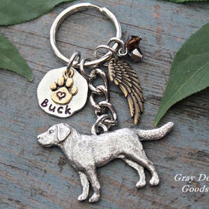 May include: Silver dog charm keyring with a paw print and the name "BUCK" engraved on it. The charm is attached to a silver chain and a silver key ring. A small gold angel wing charm is also attached to the chain. The keyring is on a wooden surface.
