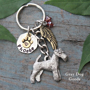 May include: A silver and gold charm keychain with a paw print charm, a heart charm, a small angel wing charm, and a silver wirehaired terrier charm. The keychain is on a wooden surface with green leaves. The charm is engraved with the name "Phoebe".