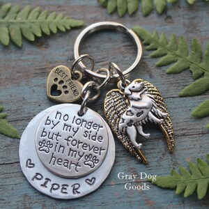 May include: Silver keychain with a round charm that reads "no longer by my side but forever in my heart" and the name "PIPER". A small heart charm with a paw print is attached to the keychain, along with a silver dog with gold wings charm. The keychain is on a wooden surface with green leaves.