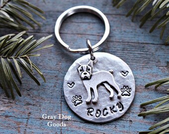 Può includere: Portachiavi con targhetta in argento con una silhouette di Boston Terrier, impronte di zampe, cuori e il nome "Rocky" inciso su di essa. La targhetta è attaccata a un portachiavi in argento. "Gray Dog Goods" è stampato sotto la targhetta.