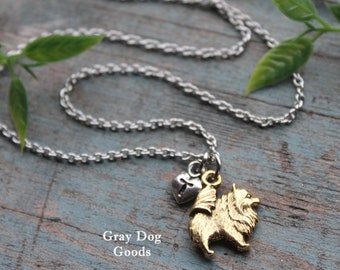May include: A silver-toned chain necklace with a gold-colored Pomeranian dog pendant and a small heart charm. The pendant is detailed with textured fur. The necklace is displayed on a blue-gray wooden surface with a sprig of green leaves. Text on the image reads "Gray Dog Goods."