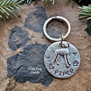 May include: Silver dog tag keychain with a silhouette of a dog, two paw prints, two hearts, and the name "Piper" engraved on it.