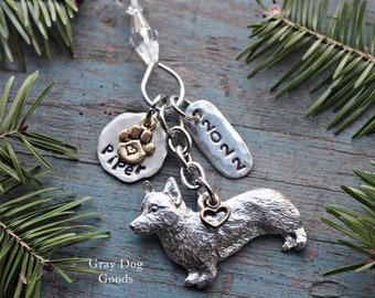 May include: A silver corgi charm keyring with a paw print charm that says "Piper" and a charm with the year "2022". The keyring has a clear crystal bead and is surrounded by green pine needles.