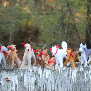 Pu&ograve; includere: Una collezione di ornamenti per cani in feltro di vari colori e razze, tra cui un levriero, un husky e un cane bianco. Gli ornamenti sono disposti su un tronco di betulla con accenti di nastro rosso.