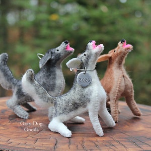 May include: Three gray and brown felt dog ornaments with pink noses, standing on a wooden surface. The middle dog has a silver tag with the year 2023 on it.