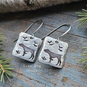 May include: A pair of square silver earrings with a dog silhouette, birds, and grass details. The earrings have a hammered texture and long, curved ear wires. The text "Gray Dog Goods" is visible at the bottom.