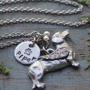 May include: Silver necklace with a charm of a dachshund with wings and a paw print charm with the name "Piper" engraved on it.