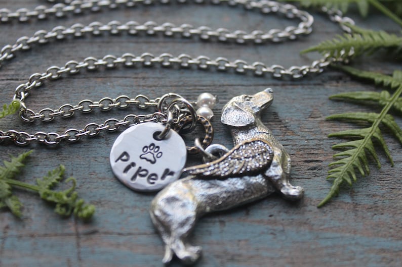 May include: Silver necklace with a charm of a dachshund with wings and a paw print charm with the name "Piper" engraved on it.