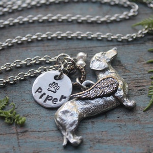 May include: Silver necklace with a charm of a dachshund with wings and a round charm with the name "Piper" and a paw print.