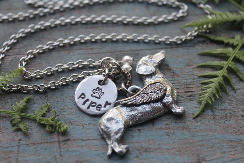 May include: Silver necklace with a charm of a dachshund with wings and a round charm with the name "Piper" and a paw print.