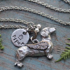 May include: Silver necklace with a dachshund charm, a paw print charm with the name "Piper" engraved on it, and a silver chain. The dachshund charm has wings and is on a wooden surface.