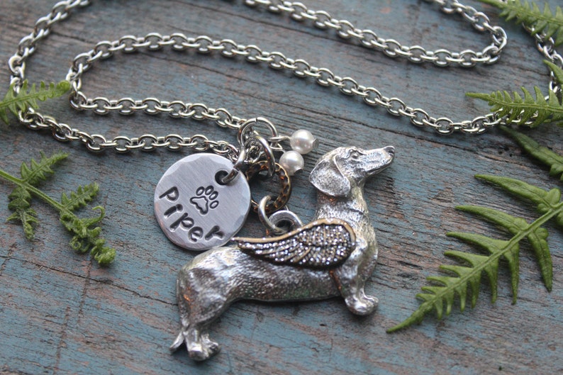 May include: Silver necklace with a dachshund charm, a paw print charm with the name "Piper" engraved on it, and a silver chain. The dachshund charm has wings and is on a wooden surface.
