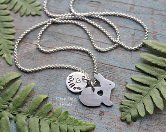 Può includere: Una collana d'argento con un ciondolo a forma di coniglio e un ciondolo rotondo con il nome "Willow" inciso. La collana è su una superficie di legno con foglie verdi.