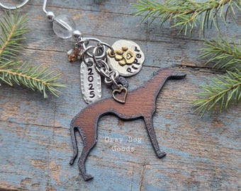 May include: A brown wooden greyhound dog keychain with a metal keyring, a heart charm, and a tag that says "2025". There is also a charm with a paw print and the name "Piper". The text "Gray Dog Goods" is visible.