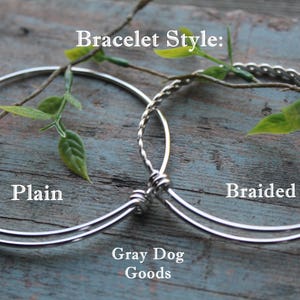 K&ouml;nnte beinhalten: Zwei silberne Armb&auml;nder, eines schlicht und eines geflochten, auf einer Holzoberfl&auml;che. Die Armb&auml;nder sind beide dehnbar und haben einen silbernen Verschluss. Der Text "Bracelet Style:" befindet sich &uuml;ber den Armb&auml;ndern, und der Text "Plain" und "Braided" befindet sich unter den Armb&auml;ndern. Der Text "Gray Dog Goods" befindet sich unter den Armb&auml;ndern.