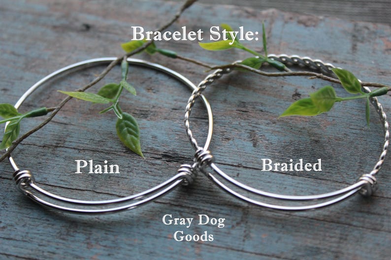 K&ouml;nnte beinhalten: Zwei silberne Armb&auml;nder, eines schlicht und eines geflochten, auf einer Holzoberfl&auml;che. Die Armb&auml;nder sind beide dehnbar und haben einen silbernen Verschluss. Der Text "Bracelet Style:" befindet sich &uuml;ber den Armb&auml;ndern, und der Text "Plain" und "Braided" befindet sich unter den Armb&auml;ndern. Der Text "Gray Dog Goods" befindet sich unter den Armb&auml;ndern.