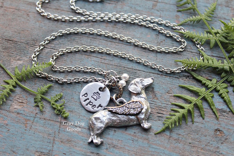 May include: Silver necklace with a dachshund charm with wings and a round charm with the name "Piper" and a paw print. The necklace is on a wooden surface with green ferns.