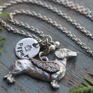 May include: Silver necklace with a dachshund charm, a round charm with the name "Piper" and a paw print, and two small pearl beads. The dachshund charm has wings.