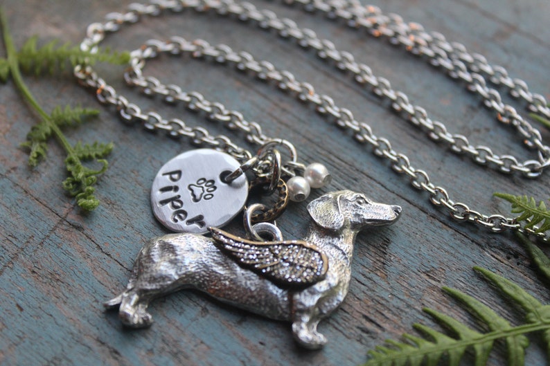 May include: Silver necklace with a dachshund charm, a round charm with the name "Piper" and a paw print, and two small pearl beads. The dachshund charm has wings.