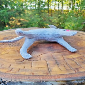 Pu&ograve; includere: Un gatto di peluche grigio in feltro sdraiato su una superficie di legno. Il gatto ha dettagli in feltro rosa sulle orecchie e sul naso.