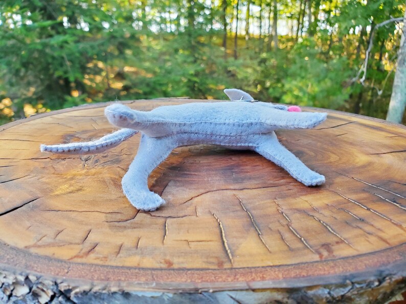 Pu&ograve; includere: Un gatto di peluche grigio in feltro sdraiato su una superficie di legno. Il gatto ha dettagli in feltro rosa sulle orecchie e sul naso.