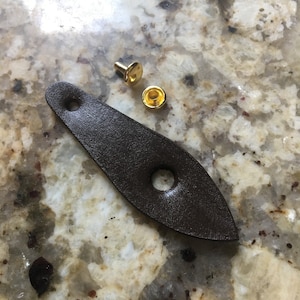 Könnte beinhalten: Ein braunes Lederband mit zwei Löchern und einem goldenen Metallnieten. Der Niet hat einen flachen Kopf und einen runden Schaft. Das Band liegt auf einer hellbraunen, gefleckten Oberfläche.