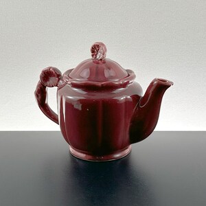 May include: A burgundy ceramic teapot with a floral design on the lid and handle. The teapot has a rounded body and a spout on the side.