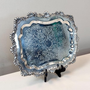 May include: A rectangular silver-toned serving tray featuring an ornate, embossed floral and scrollwork pattern across its center. The tray has a raised, scalloped edge with detailed decorative molding along the entire perimeter of the frame.