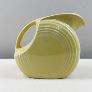以下が含まれることがあります： 淡い黄色の陶器製ピッチャー。円盤状の丸みを帯びた本体の側面には同心円状のエンボス加工が施され、上部には曲線的なハンドルが一体化しており、縁からは幅広で尖った注ぎ口が突き出ている。
