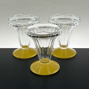 May include: Three clear glass sundae dishes with a yellow base. The dishes have a fluted, tulip-shaped bowl and a scalloped rim. The yellow base is round and provides a stable foundation for the glass.