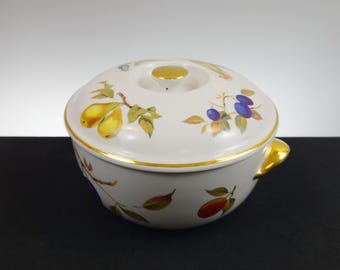 May include: A white ceramic serving bowl with a lid, adorned with a fruit design featuring yellow pears and purple plums. The bowl has a gold rim and handle, and a gold knob on the lid. A decorative kitchenware item.