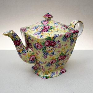 Könnte beinhalten: Eine quadratische Teekanne aus Porzellan mit einem leuchtend gelben Hintergrund, der mit einem dichten Blumenmuster aus rosa Rosen, violetten Blüten und kleinen gelben Blumen bedeckt ist. Die Kanne hat einen passenden Deckel mit Knauf und Goldrand.