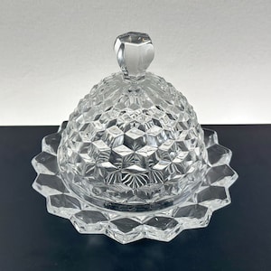 Puede incluir: Un mantequillera de vidrio transparente con tapa abovedada y base festoneada. La tapa tiene un pomo en la parte superior.