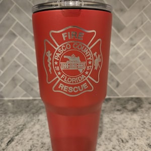 May include: A red insulated tumbler with a silver lid. The tumbler features a silver emblem with the words "FIRE PASCO COUNTY FLORIDA RESCUE" and the years "18" and "87". The tumbler is designed to keep beverages hot or cold.