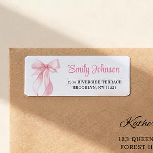 May include: A white rectangular label with a watercolor pink bow and the name "Emily Johnson" in pink script. The address "1234 RIVERSIDE TERRACE BROOKLYN, NY 11231" is printed below. The label is on a brown envelope.