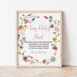 May include: A printable sign for a baby shower game. The sign has a floral wreath border and the text "Tiny Baby Hunt" in pink. The text below reads "Multiple babies have been hidden throughout the shower. Whoever finds the most at the end, wins a prize! Happy Hunting!"