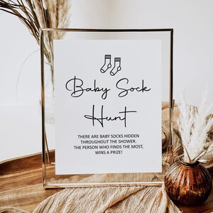 May include: A gold-framed sign with white paper inside. The sign says "Baby Sock Hunt" with a small illustration of two baby socks. The text below reads "There are baby socks hidden throughout the shower. The person who finds the most, wins a prize!"