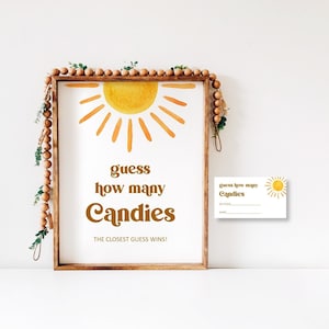 May include: A wooden sign with a yellow sun graphic and the text "guess how many candies the closest guess wins!"  A smaller card with a sun graphic and the text "guess how many candies" is below the sign.