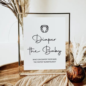 May include: A white sign with a gold frame, a black diaper icon, and the text "Diaper the Baby" in black cursive font. Below the text, it reads "Who can diaper their baby the fastest blindfolded?"