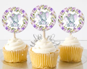 May include: Three purple and white cupcake toppers with a gray elephant and floral design. The toppers are on top of cupcakes with white frosting.