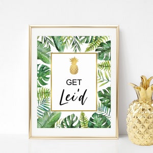 May include: A gold framed print with a white background and green tropical leaves. The print has a gold pineapple and the text "GET Lei'd".
