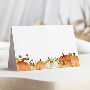 May include: A folded white card featuring a watercolor illustration along the bottom edge. The design displays a row of various pumpkins in shades of orange, white, and tan, accented with green vines, leaves, and small flowers against a plain background.