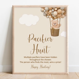 May include: A brown and beige printable sign with a teddy bear in a hot air balloon made of balloons. The sign says "Pacifier Hunt" and "Multiple pacifiers have been hidden throughout the shower. The person who finds the most, wins a prize! Happy Hunting!"