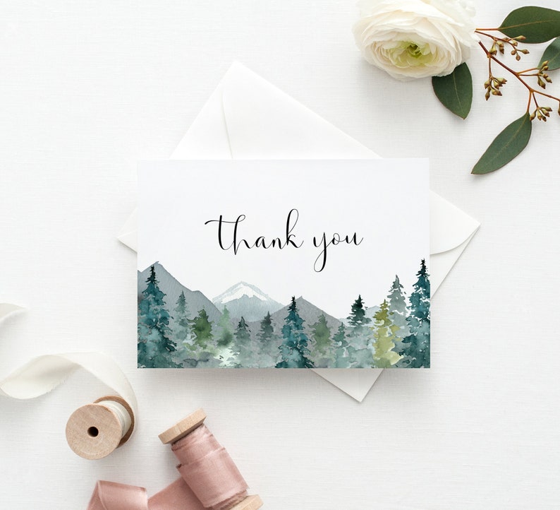 May include: A white card with the words "thank you" in elegant script. The card features a watercolor landscape of mountains and evergreen trees in shades of green and blue. An envelope and floral accents complete the scene.