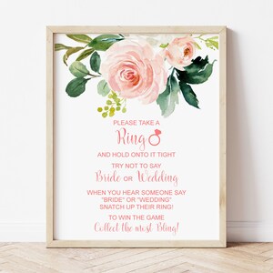 May include: A white sign with pink floral accents and the text "PLEASE TAKE A Ring AND HOLD ONTO IT TIGHT TRY NOT TO SAY Bride OR Wedding WHEN YOU HEAR SOMEONE SAY "BRIDE" OR "WEDDING" SNATCH UP THEIR RING! TO WIN THE GAME Collect the most Bling!"