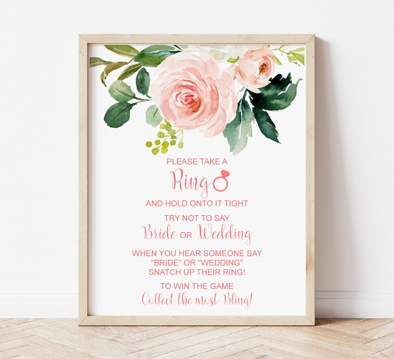 May include: A white sign with pink floral accents and the text "PLEASE TAKE A Ring AND HOLD ONTO IT TIGHT TRY NOT TO SAY Bride OR Wedding WHEN YOU HEAR SOMEONE SAY "BRIDE" OR "WEDDING" SNATCH UP THEIR RING! TO WIN THE GAME Collect the most Bling!"