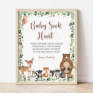 May include: A printable sign for a baby shower game. The sign has a brown wooden frame and a white background with green foliage around the edges. The text on the sign reads "Baby Sock Hunt" and includes instructions for the game. The sign also features illustrations of woodland animals, including a deer, a fox, a raccoon, and a bear.
