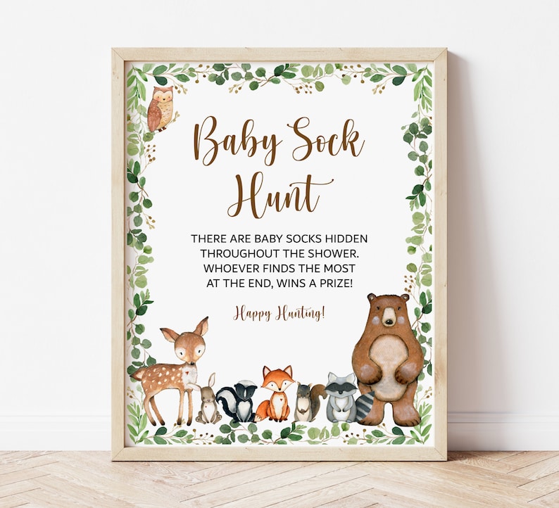 May include: A printable sign for a baby shower game. The sign has a brown wooden frame and a white background with green foliage around the edges. The text on the sign reads "Baby Sock Hunt" and includes instructions for the game. The sign also features illustrations of woodland animals, including a deer, a fox, a raccoon, and a bear.