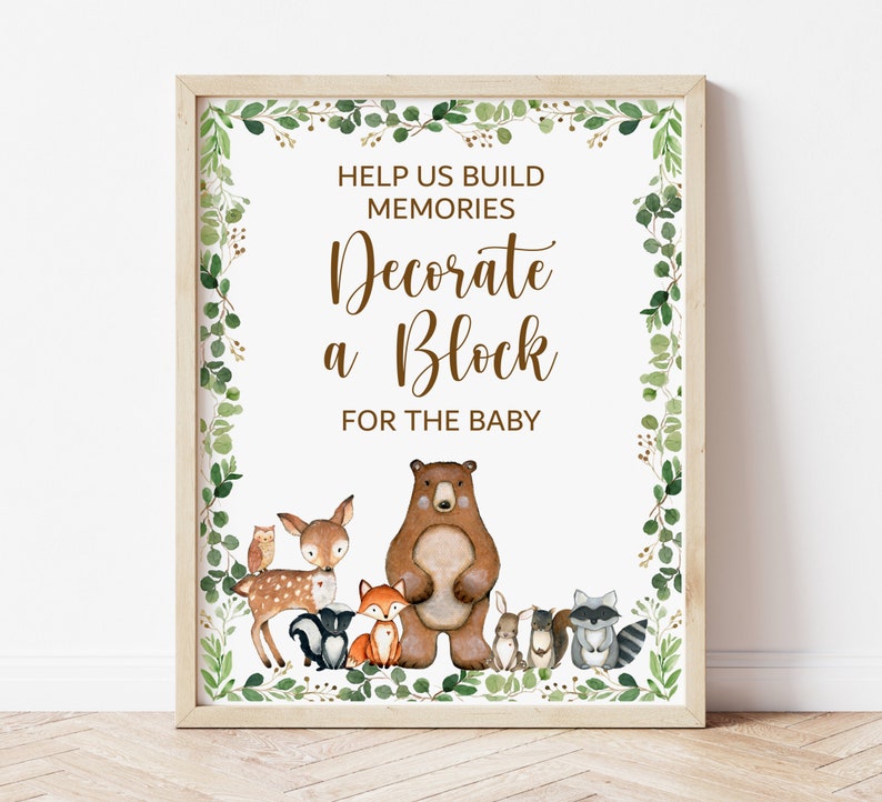 May include: A white sign with a brown border and green foliage around the edges. The sign says "Help us build memories Decorate a Block for the baby". There are woodland animals on the sign, including a bear, deer, fox, owl, raccoon, and squirrel.